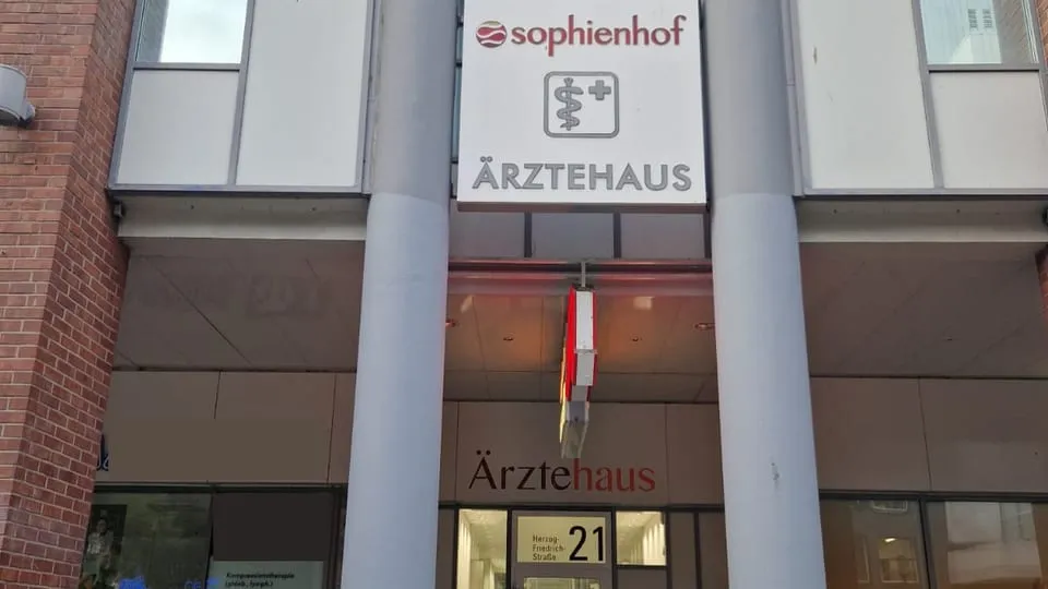 Eingang des Ärztehauses Sophienhof zwischen zwei Säulen: darüber das Schild „sophienhof Ärztehaus“, darunter der Schriftzug „Ärztehaus“ und die Glastür mit der Hausnummer 21.