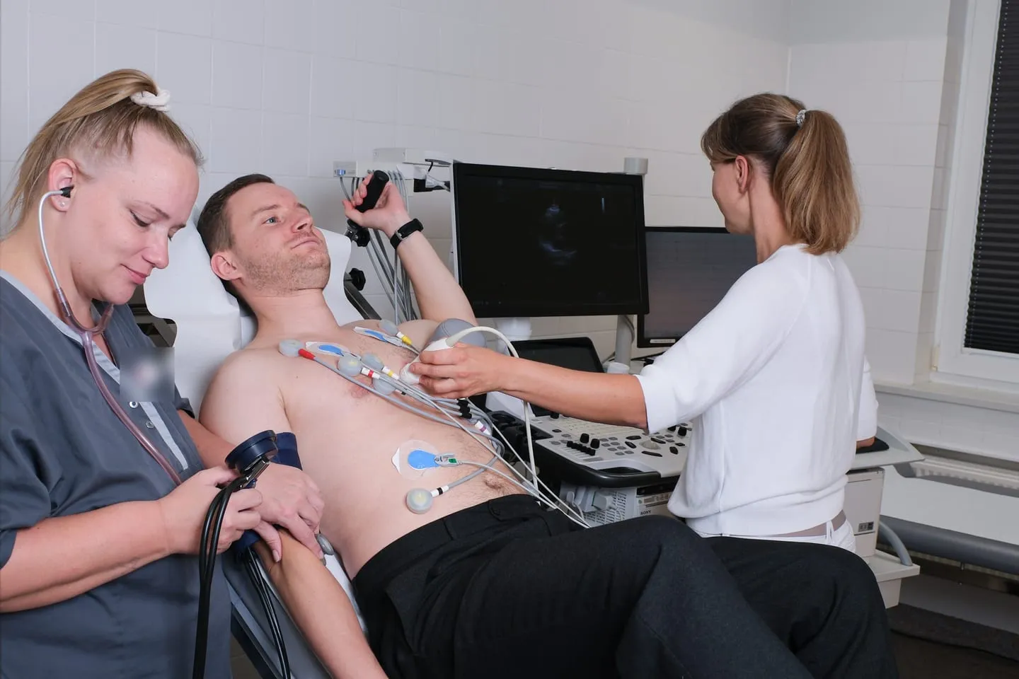 Stressechokardiographie: Ein Patient mit EKG-Elektroden hält halb liegend den Haltegriff der Liege, eine Medizinische Fachangestellte misst mit Stethoskop den Blutdruck, eine Ärztin untersucht das Herz mit Ultraschall.