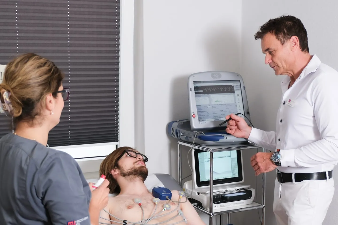 Kontrolle eines Herzschrittmachers: Ein Arzt liest das Gerät mit dem Programmiergerät aus, eine Medizinische Fachangestellte betreut den liegenden Patienten.