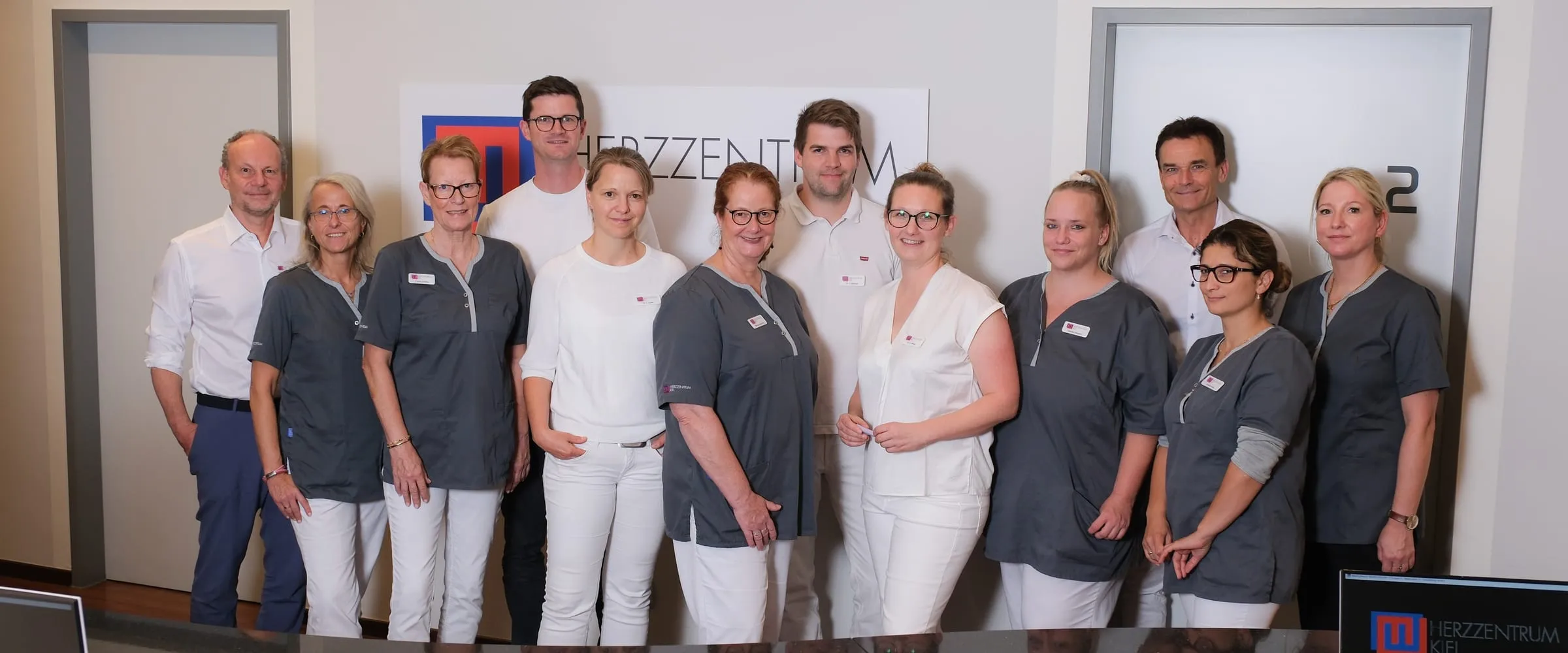 Zwölf Ärztinnen, Ärzte und Medizinische Fachangestellte stehen nebeneinander vor dem Praxisschild des Herzzentrums Kiel.